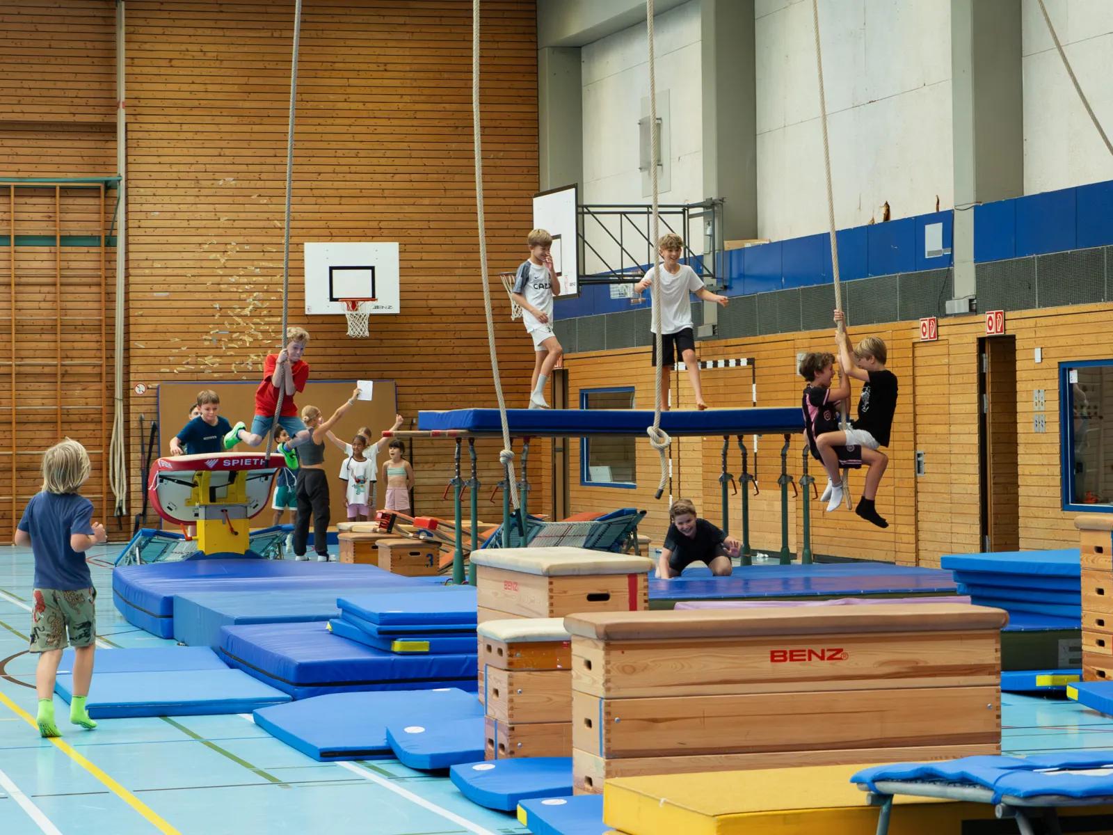 Gruppe jugendlicher Trainierender sitzt lächelnd auf einer Matte in der Sporthalle vor einer Kletterwand