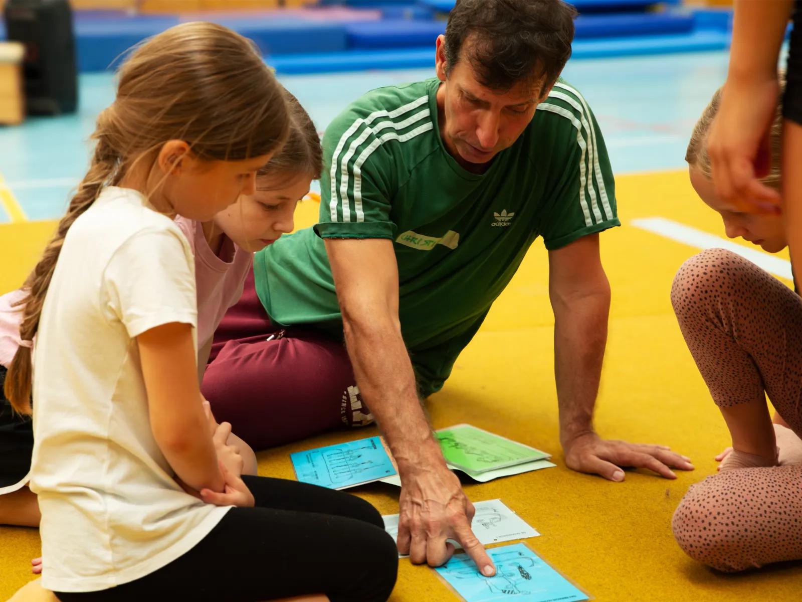 Trainer sitzt mit Kindern auf gelber Matte und erklärt anhand eines Lehrblatts eine Bewegung